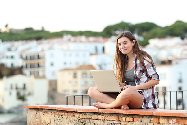 Jeune fille brune assise sur le rebord de son balcon et passant un achat sur un site e-commerce